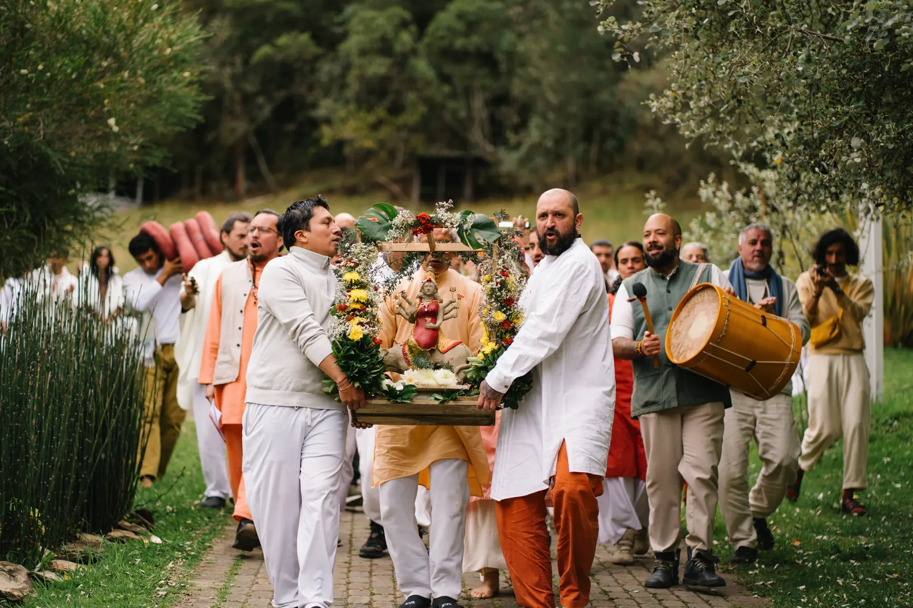 Procesión por el camino del ashram: dos hombres cargan una estatua adornada con flores, con un tambor y más personas detrás