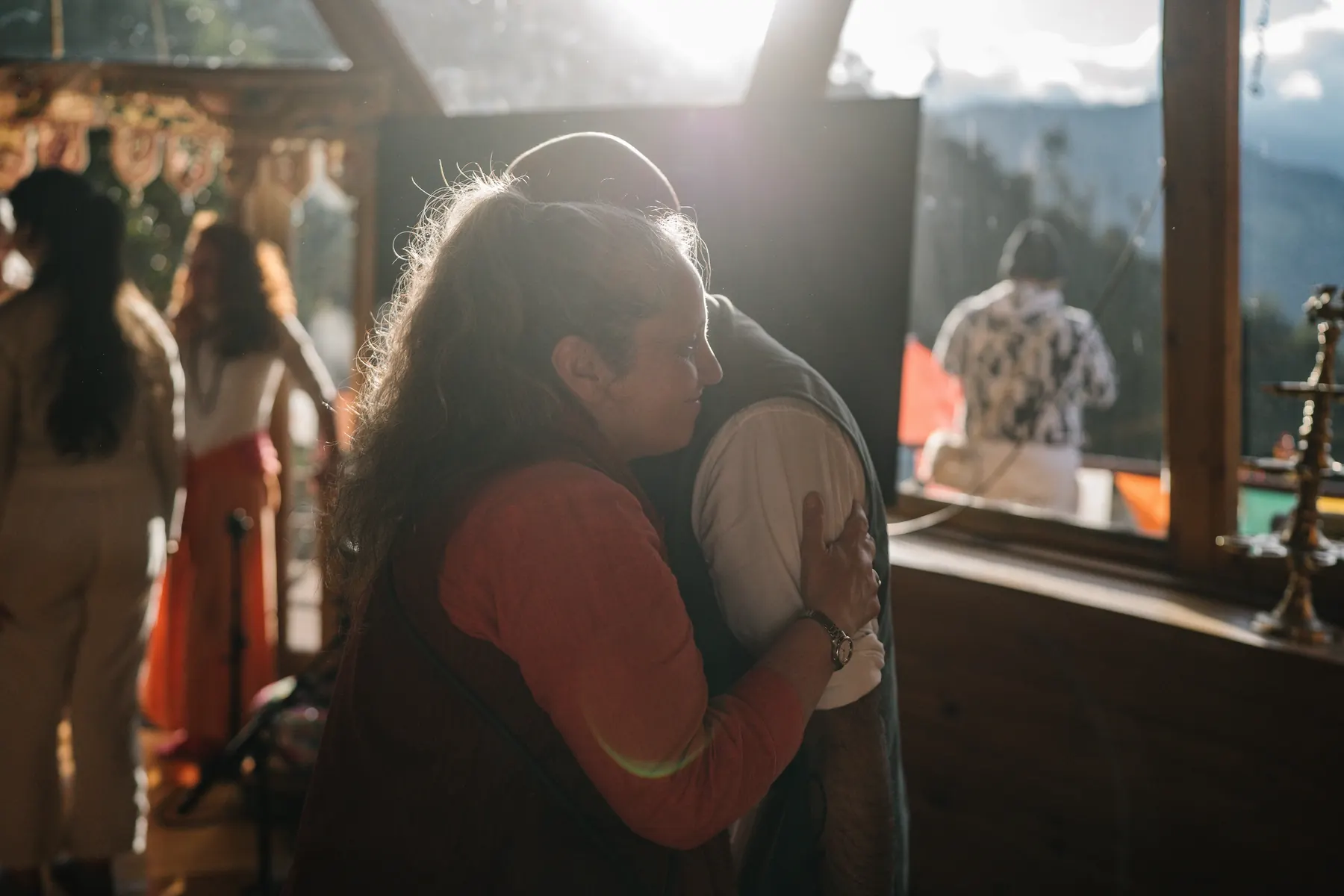Dos personas se abrazan a contraluz junto a una ventana, con la montaña al fondo