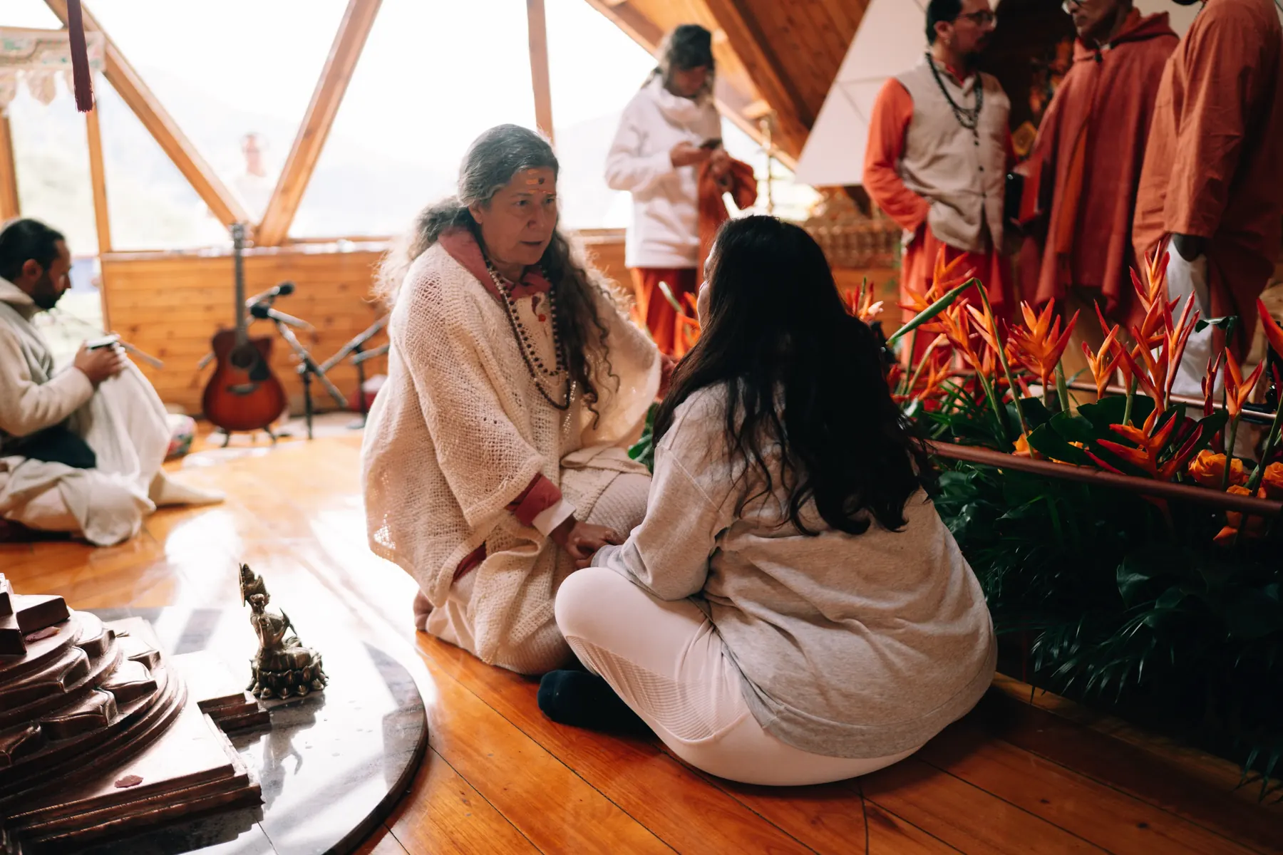 Dos personas sentadas en el suelo del templo conversan en voz baja, junto a flores naranjas
