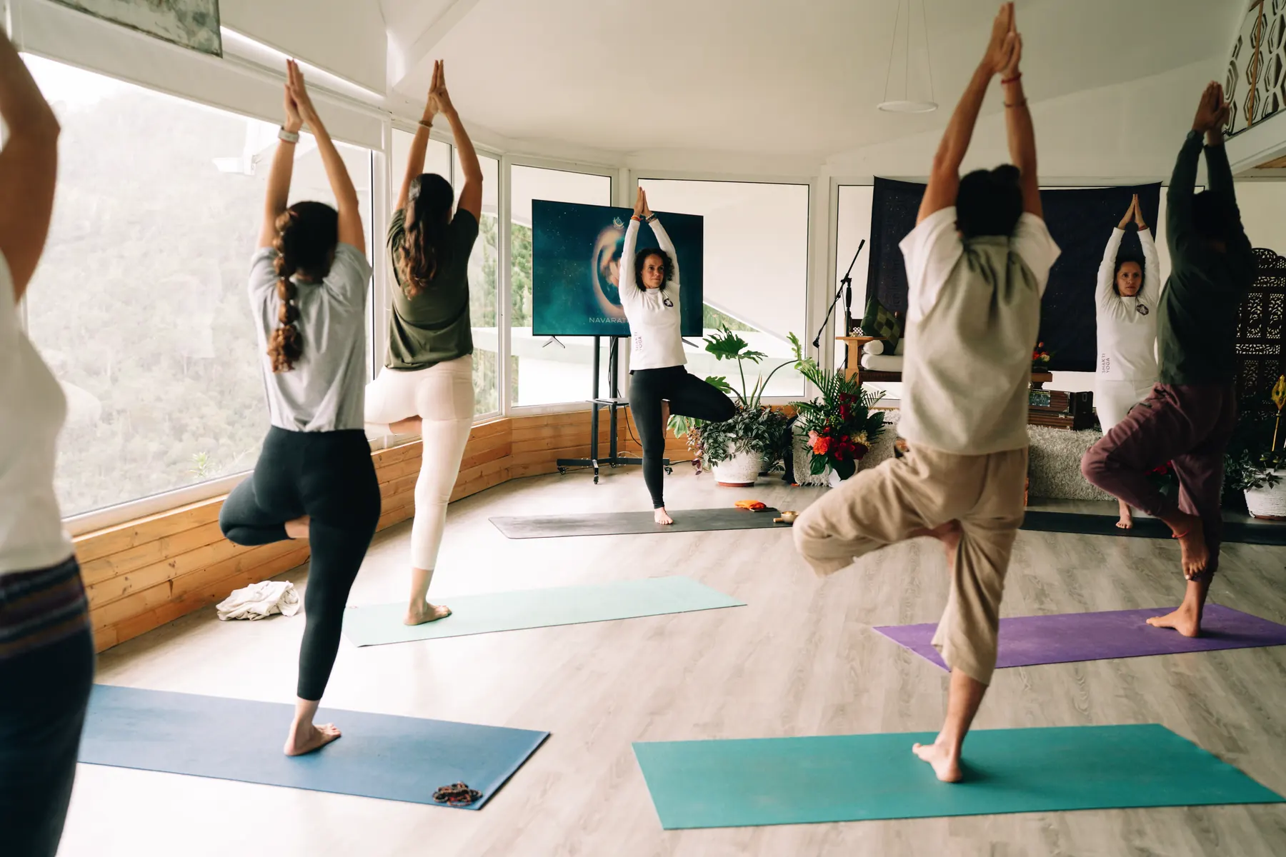 Practicantes de yoga en postura de árbol, con los brazos en alto, en una sala con ventanales