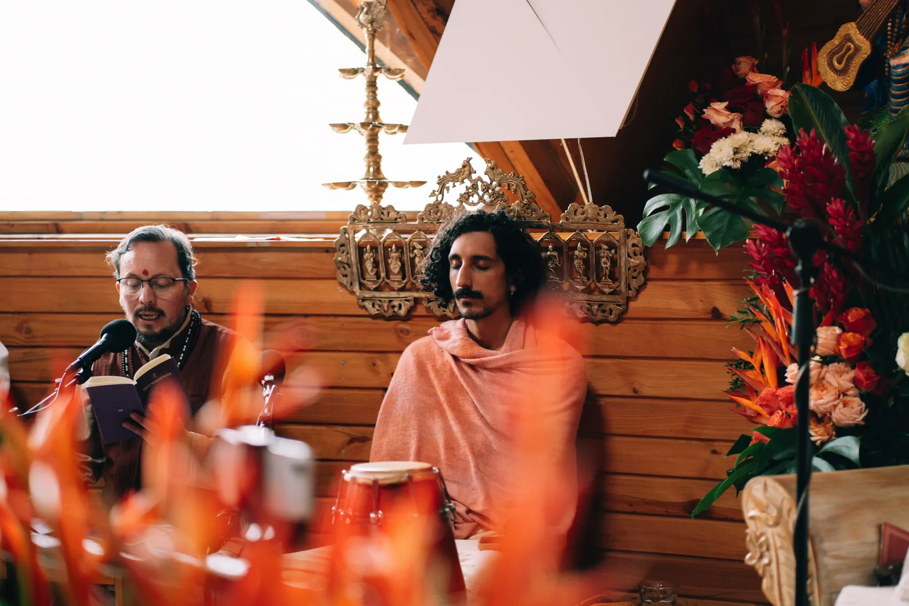 Dos hombres sentados junto al altar: uno lee con un micrófono y el otro, con un chal rosado, escucha, entre flores