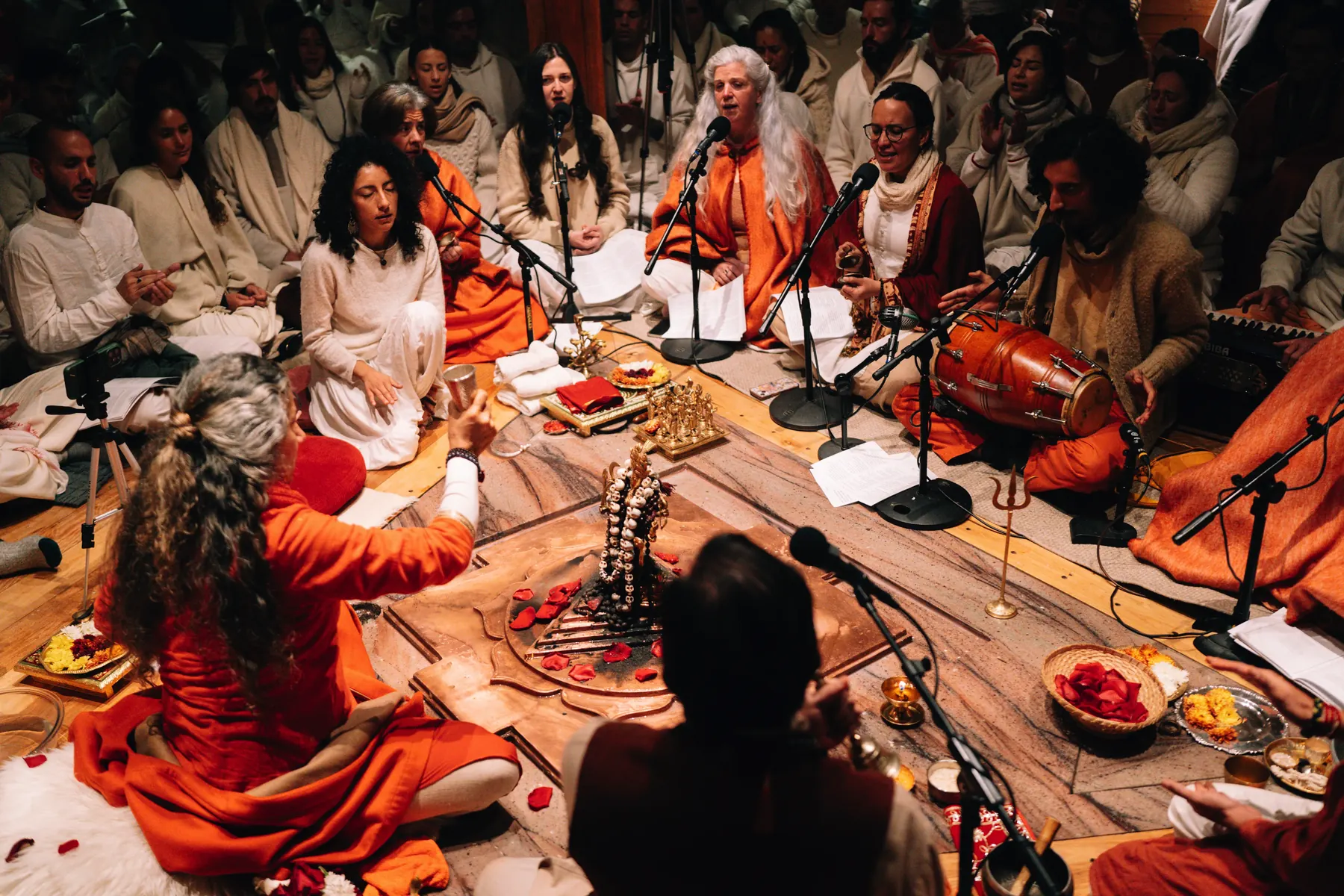 Vista desde arriba de un círculo de kirtan: músicos y cantantes en torno al altar, rodeados de participantes de blanco