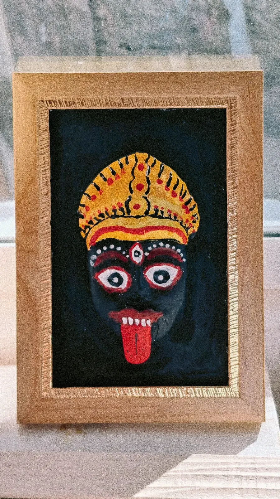 Rostro de Mahakali de piel azul oscura, con lengua roja y corona dorada, pintado a mano sobre fondo negro en un marco de madera