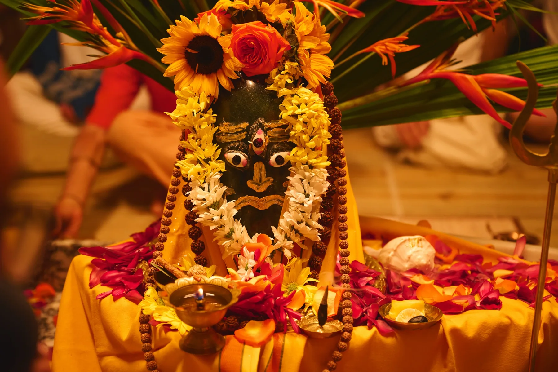 Lingam de piedra oscura con ojos y marcas de pasta amarilla, coronado de girasoles y una rosa, rodeado de guirnaldas de flores, rudrakshas, pétalos y una lámpara encendida
