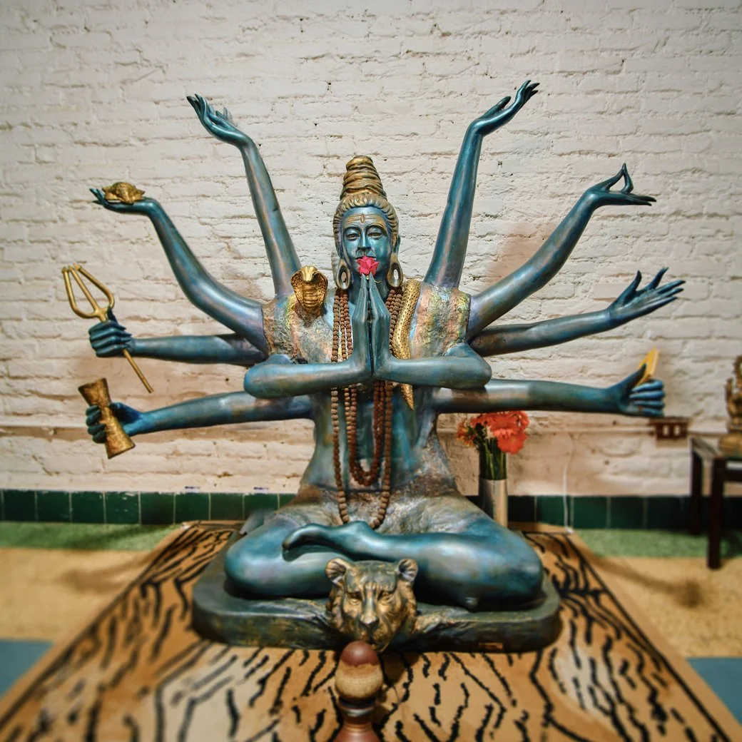 Escultura azul del Señor Shiva con varios brazos, sentado en meditación sobre una piel de tigre, con tridente, tambor damaru y rosario, ante una pared de ladrillo blanco