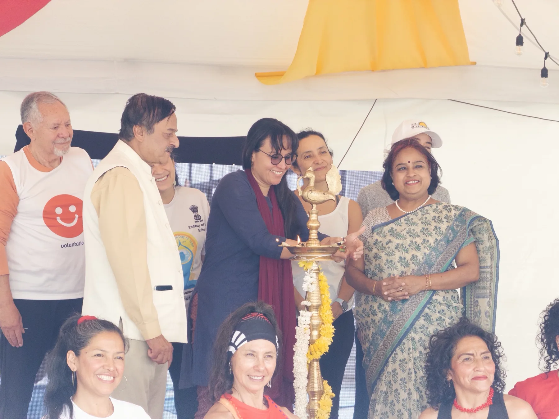 Una mujer con bufanda granate enciende una lámpara de bronce adornada con flores amarillas y blancas, rodeada de personas sonrientes, junto a una mujer con sari