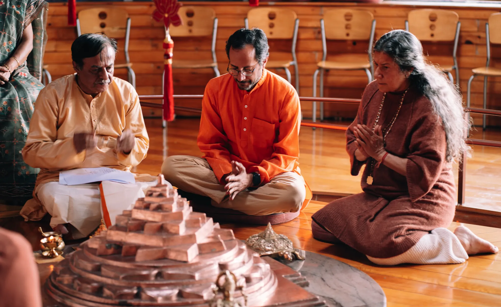 Tres personas sentadas en el suelo del templo, una de ellas con las manos en oración, frente a un yantra de cobre
