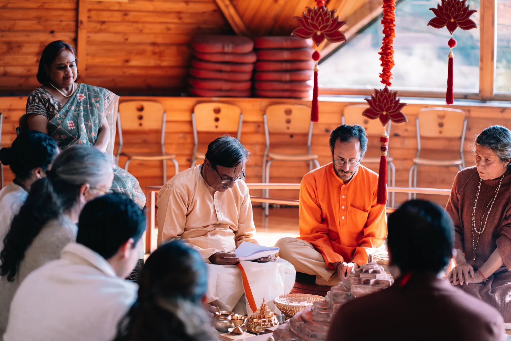 Un hombre con kurta color crema lee un texto sentado frente a un yantra de cobre, junto a otras personas reunidas en el templo
